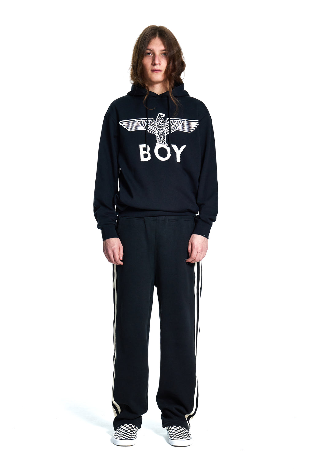 Person wearing a black hoodie with 'BOY' London logo and black pants with white stripes on a white background