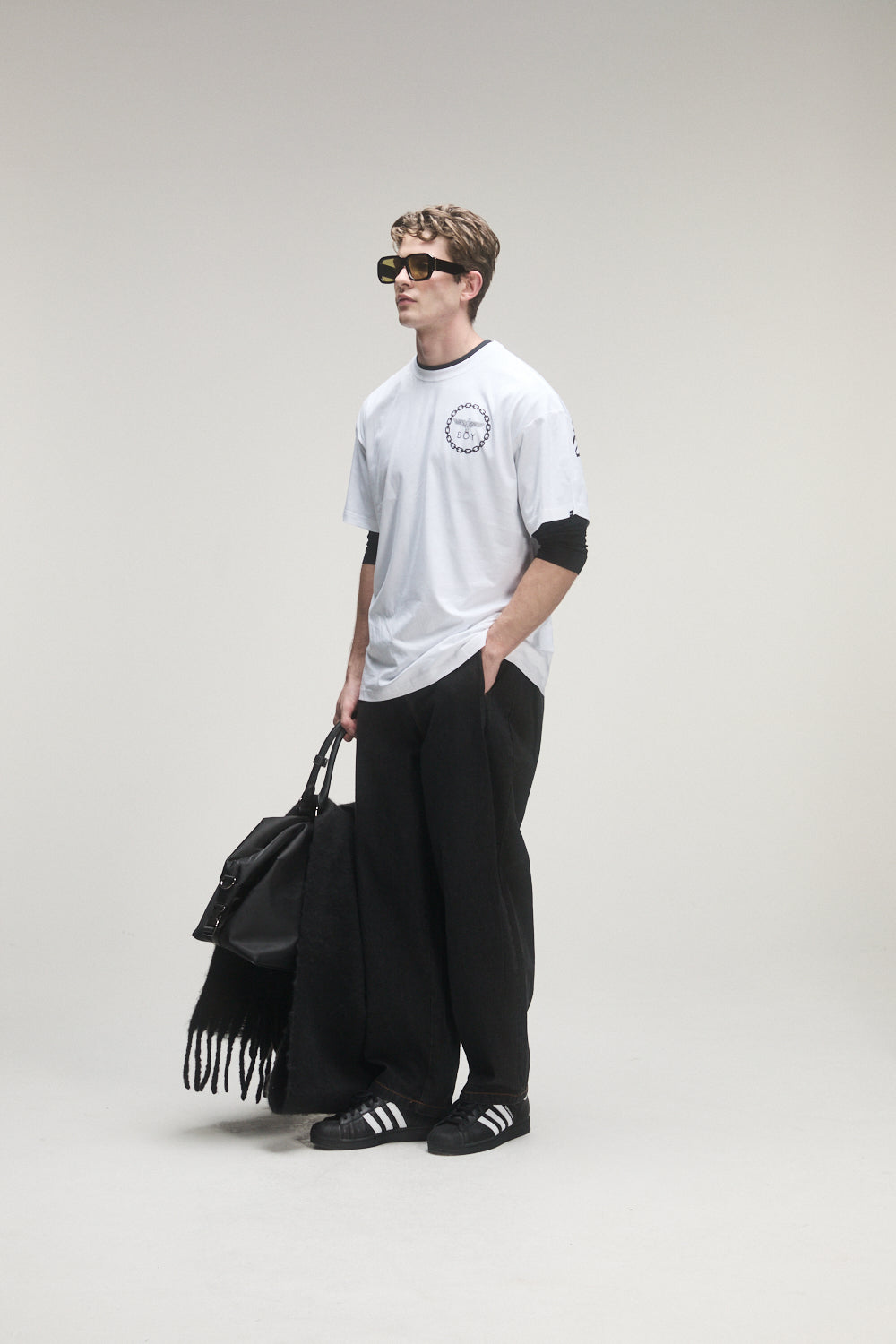 Model wearing a white BOY London t-shirt, black pants, and carrying a black bag on a plain background