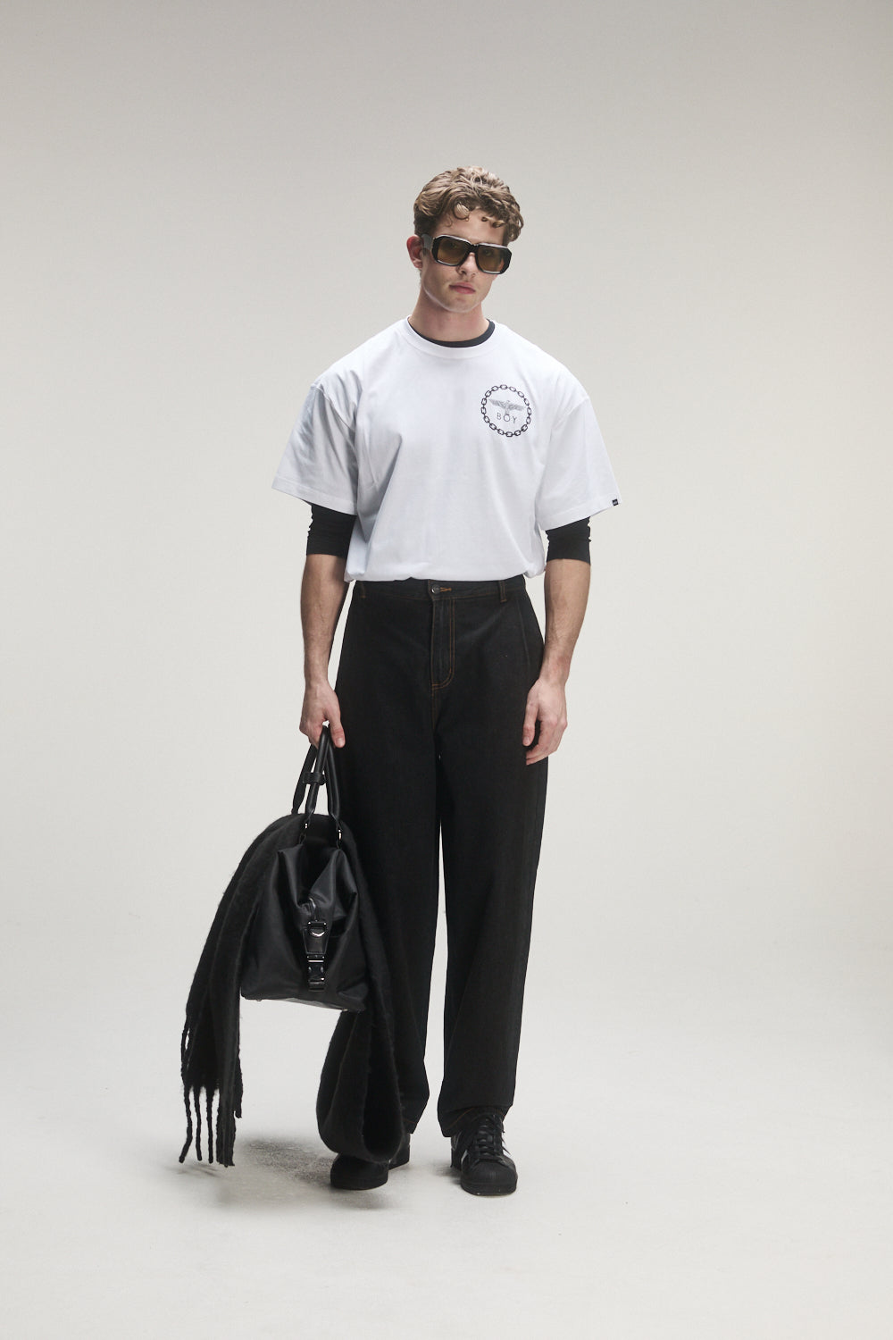 Model wearing a BOY London white t-shirt with black sleeves and black pants, holding a black bag on a light gray background