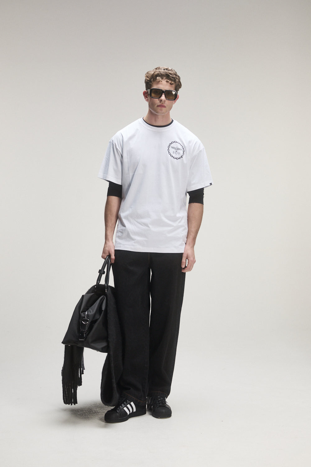 Model wearing a white BOY London t-shirt with black sleeves and pants, holding a black bag on a gray background