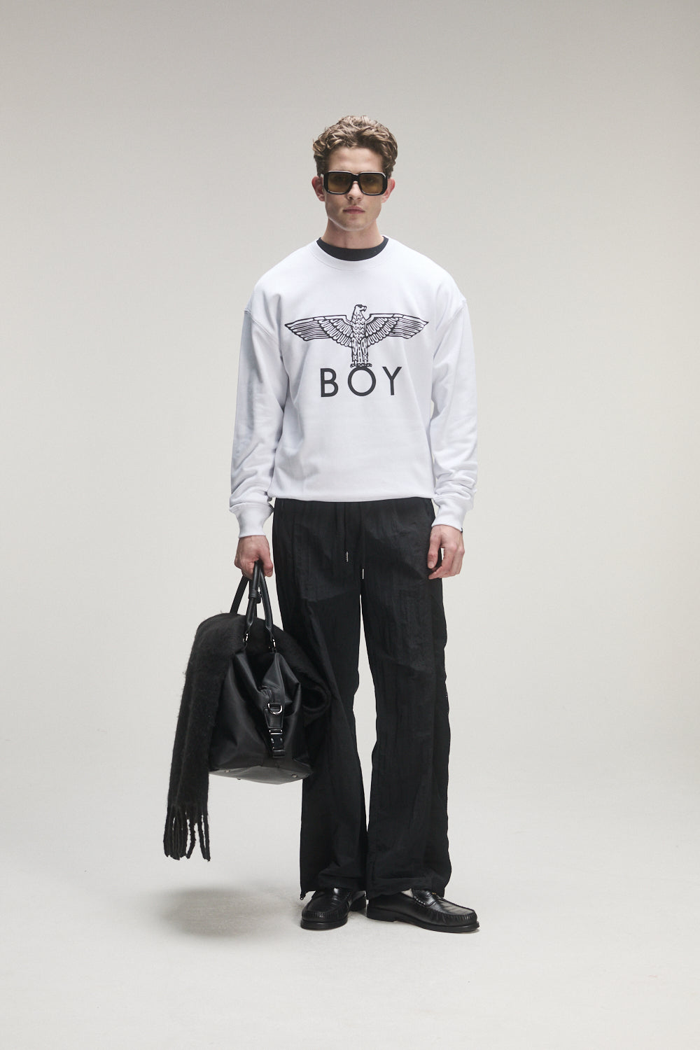 Man wearing a white sweatshirt with 'BOY'  London logo, holding a black bag on a plain background