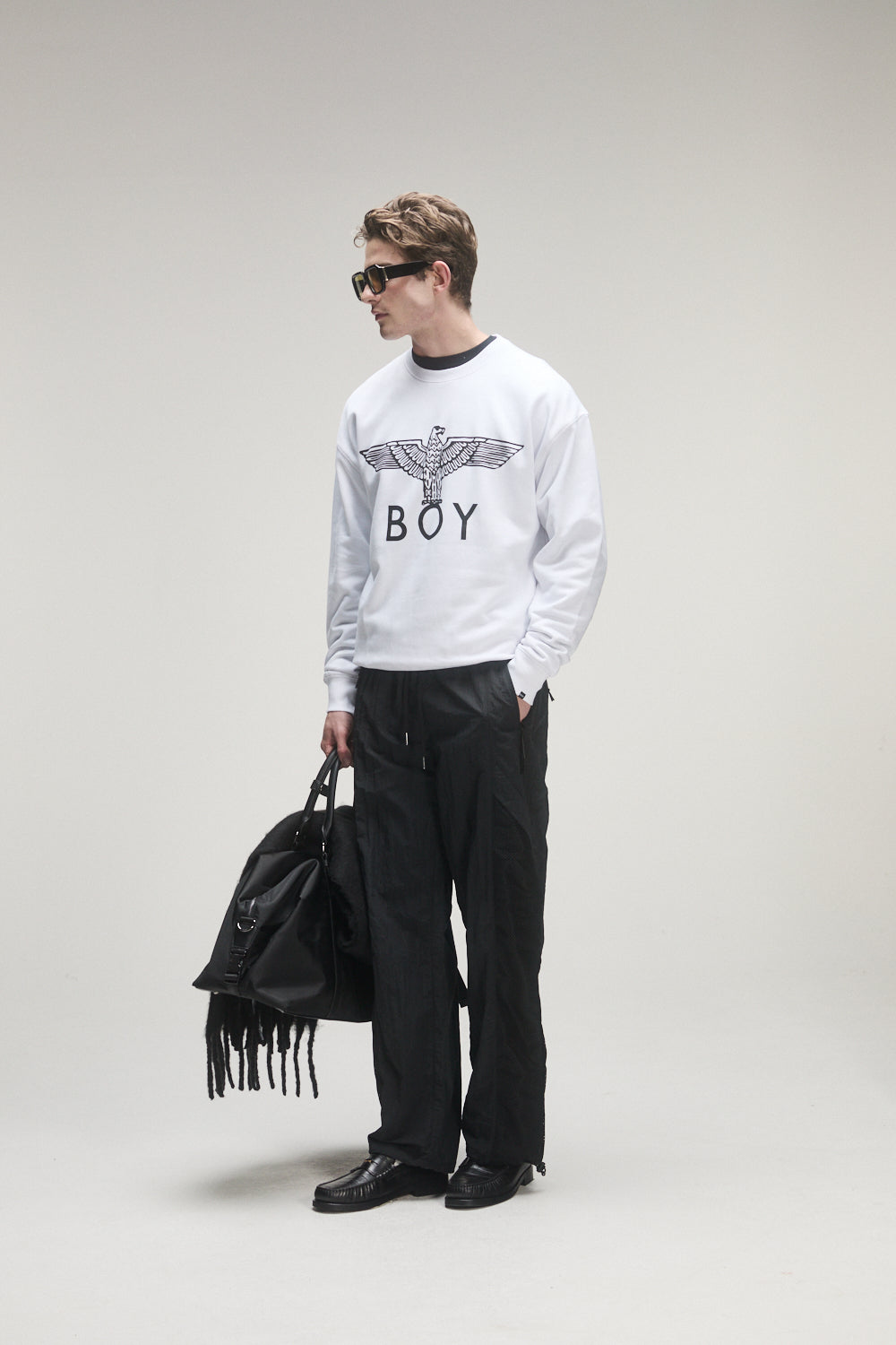 Person wearing a white sweatshirt with 'BOY' London logo, black pants, and holding a black bag on a light gray background