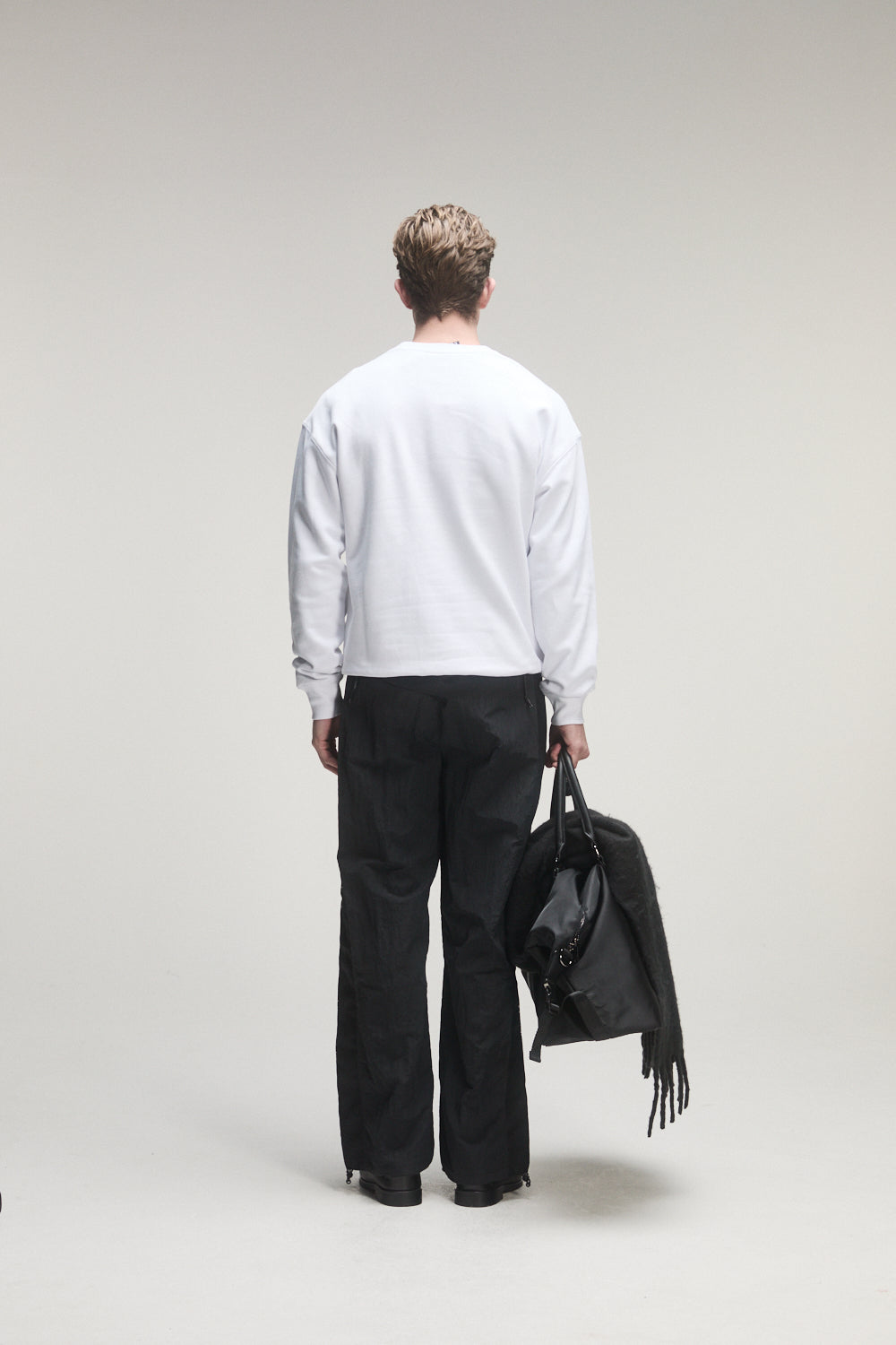 Person wearing a white BOY London sweater and black pants holding a black jacket on a plain background