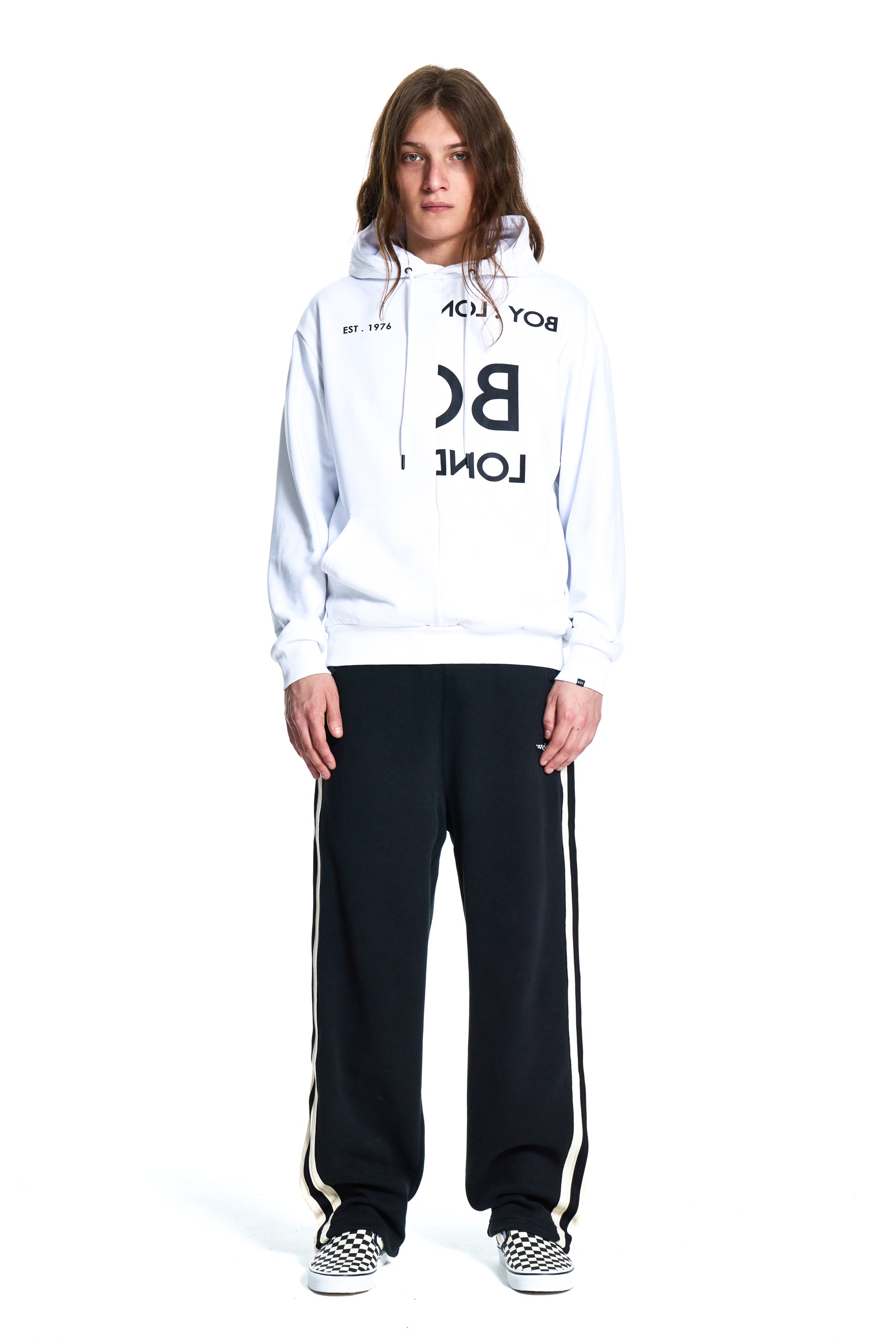 Person wearing a white hoodie with black text and black pants with white stripes on a white background