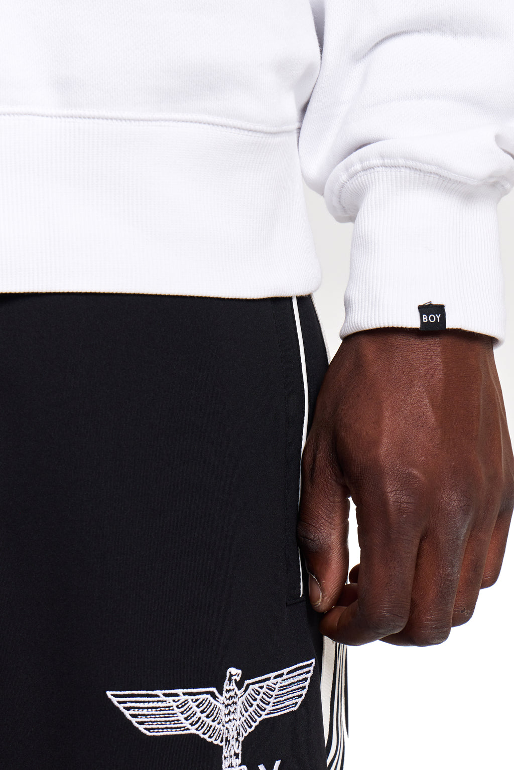 Model wearing a white BOY London sweatshirt with a brand logo, black pants with an eagle emblem