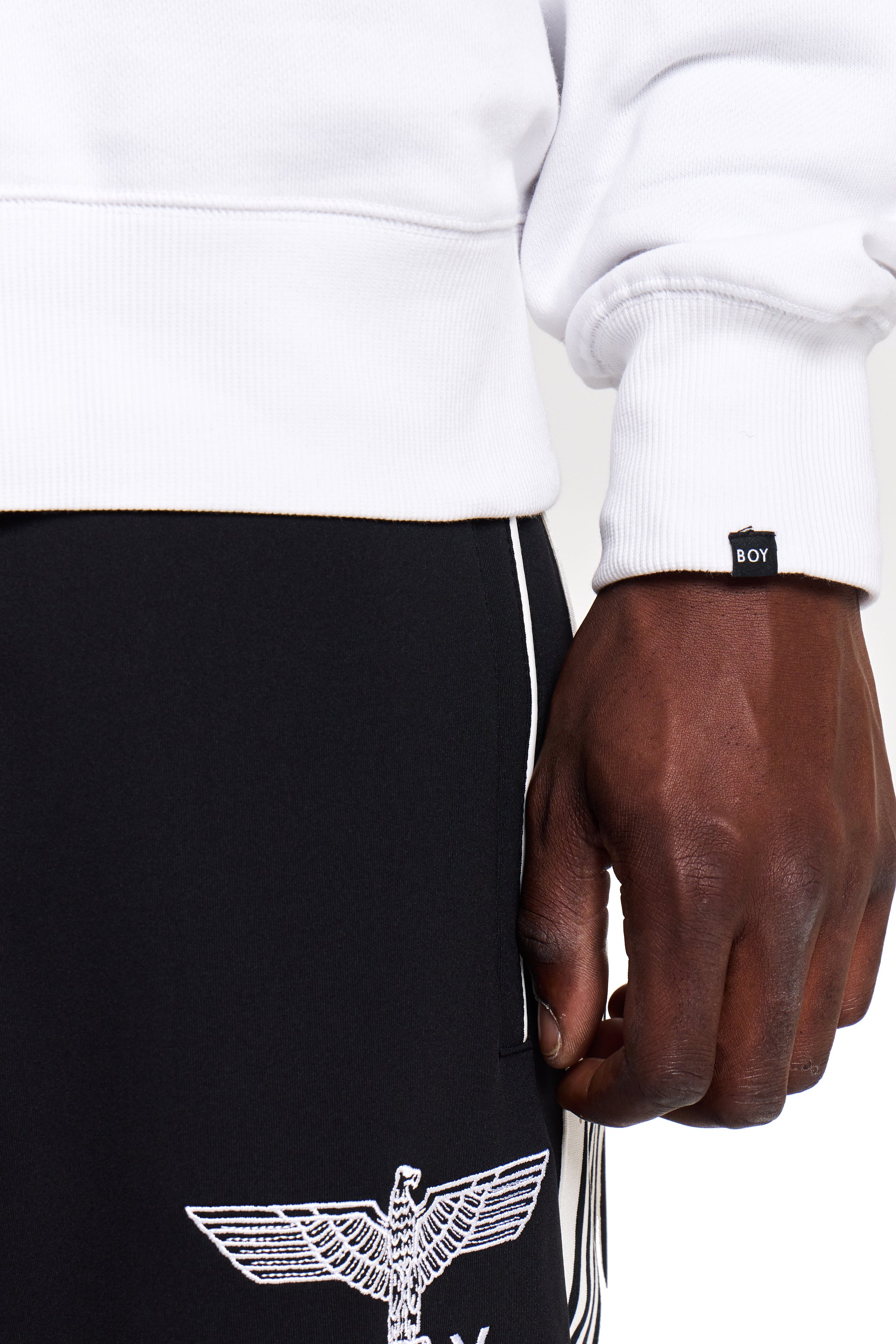 Model wearing a white BOY London sweatshirt with a brand logo, black pants with an eagle emblem