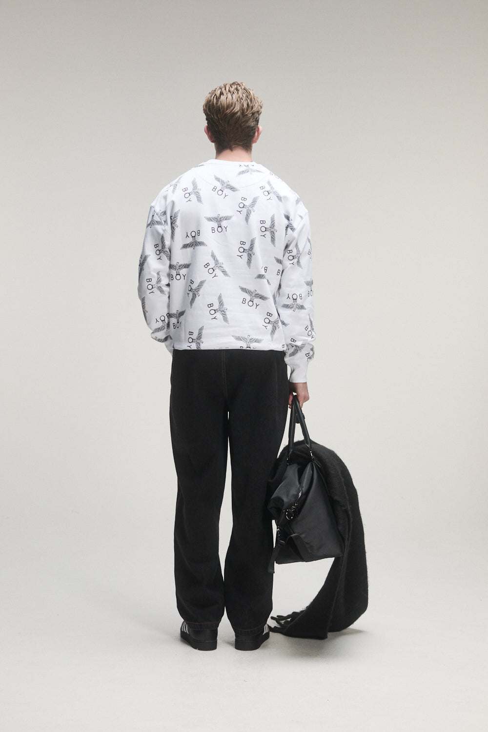 Person wearing a Boy London white patterned shirt and black pants holding a black jacket on a plain background