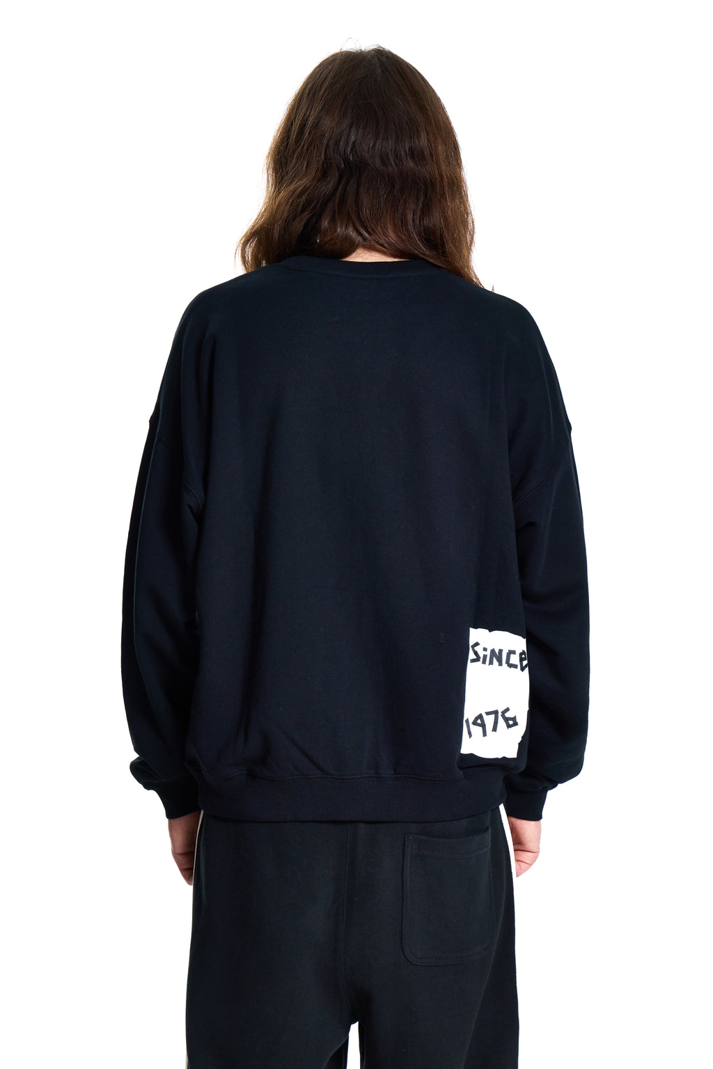 Person wearing a black BOY London sweatshirt with 'SINCE 1976' text on the sleeve, standing against a white background.