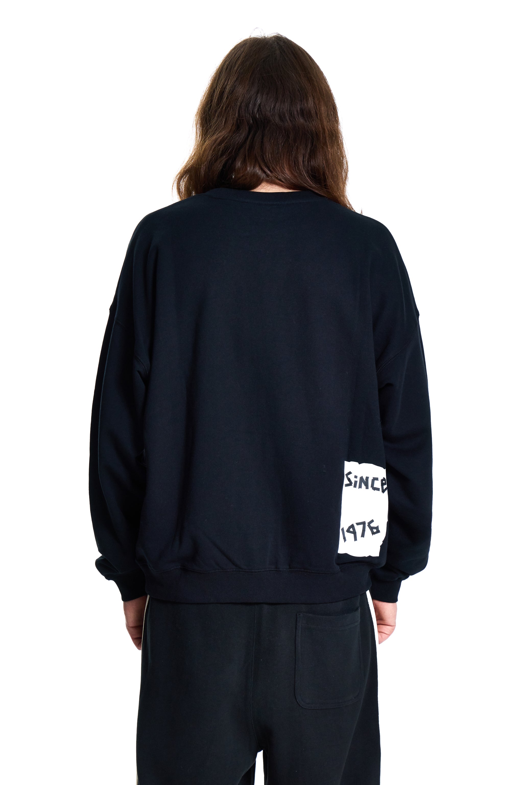 Person wearing a black sweatshirt with 'SINCE 1976' text on the sleeve, standing against a white background.