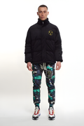 Man wearing a black BOY London jacket with a logo and colorful pants on a white background