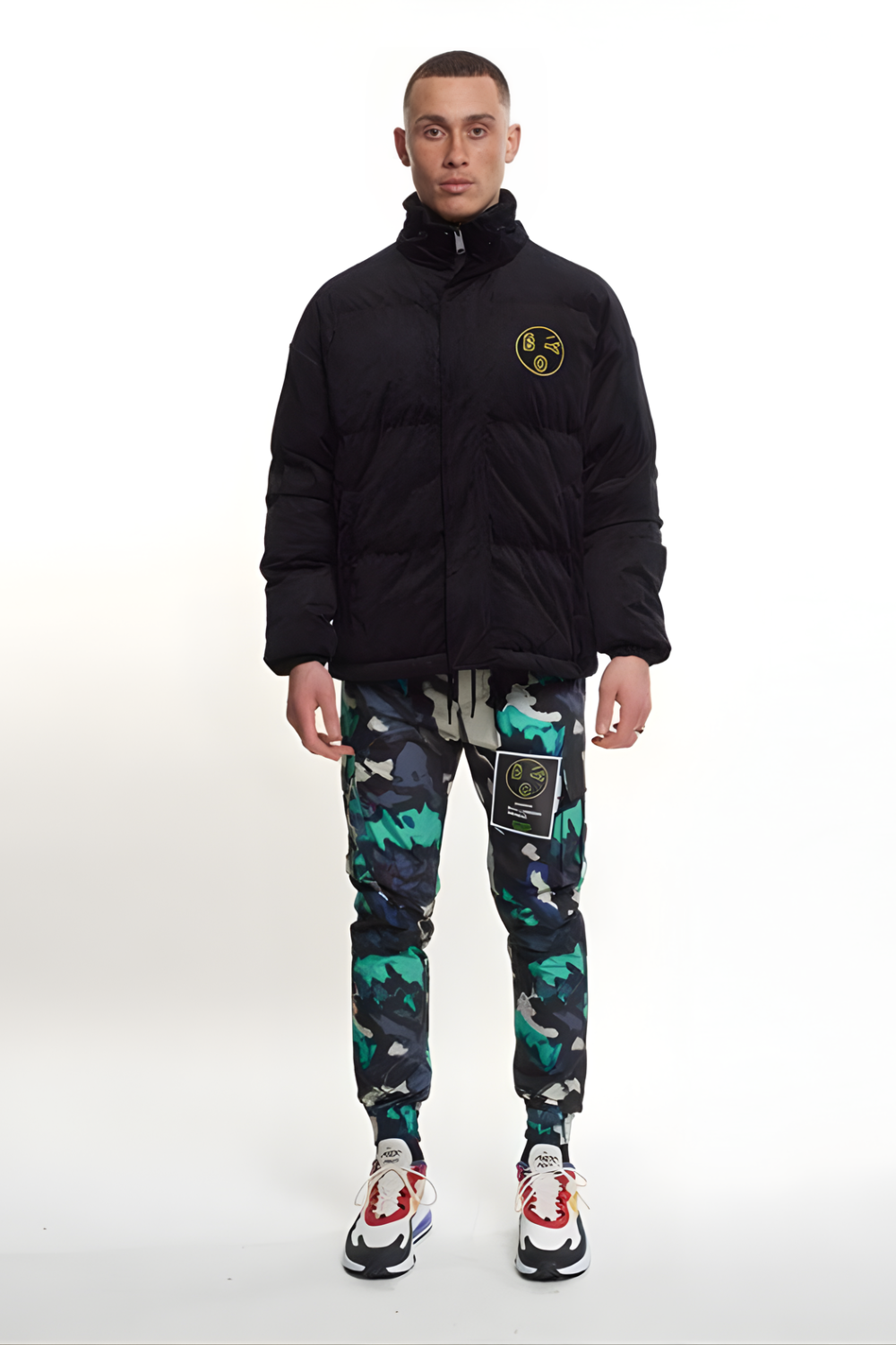 Man wearing a black BOY London jacket with a logo and colorful pants on a white background