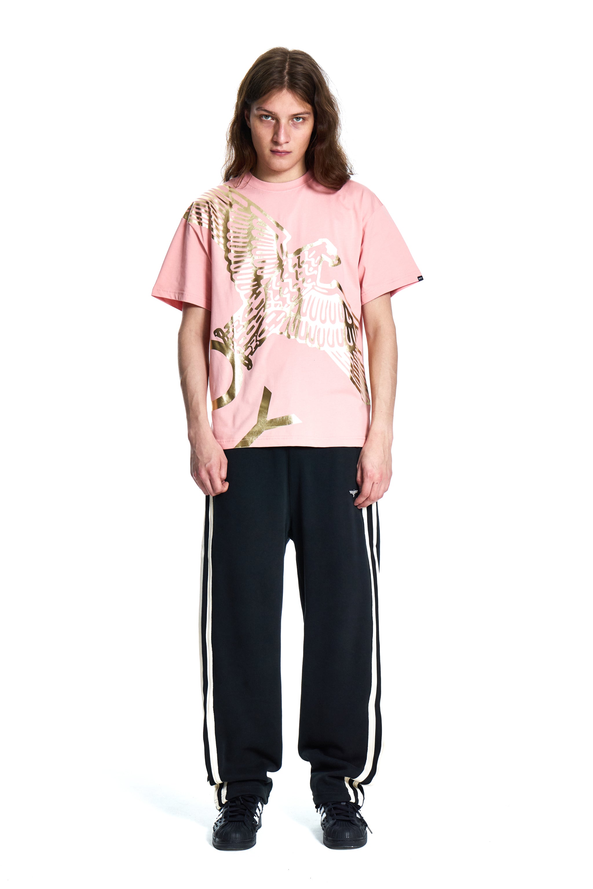Person wearing a pink BOY LONDON t-shirt with a graphic design and black pants with white stripes on a white background