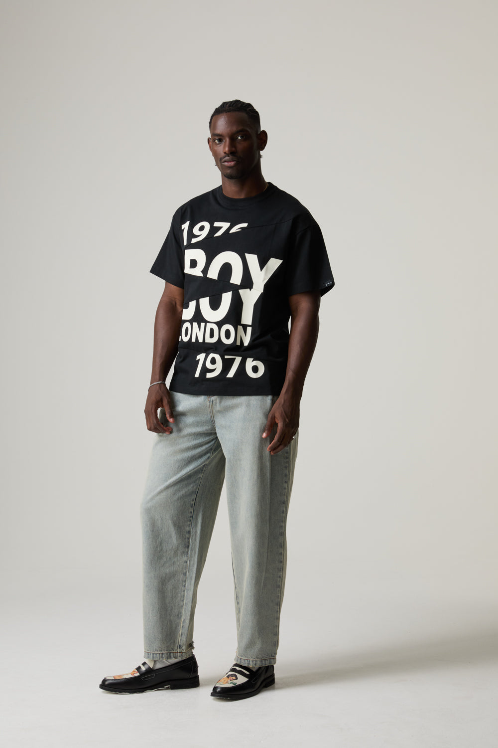 Model wearing a BOY LONDON black t-shirt with text and light gray pants on a white background