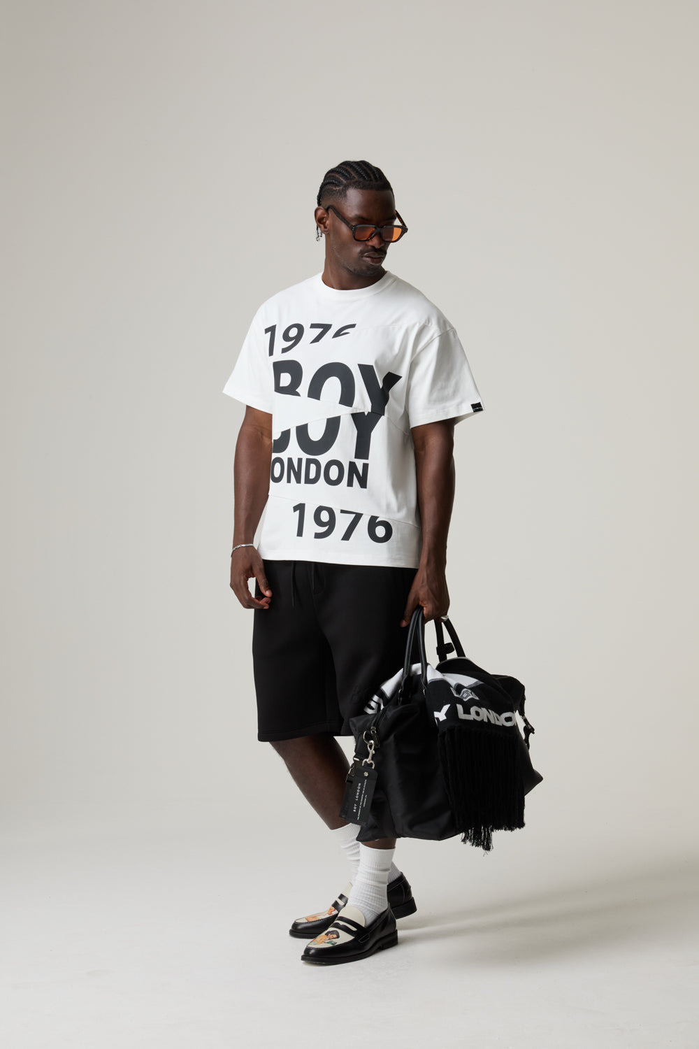Model wearing a white 'BOY LONDON' t-shirt and black shorts, holding a matching bag on a plain background.