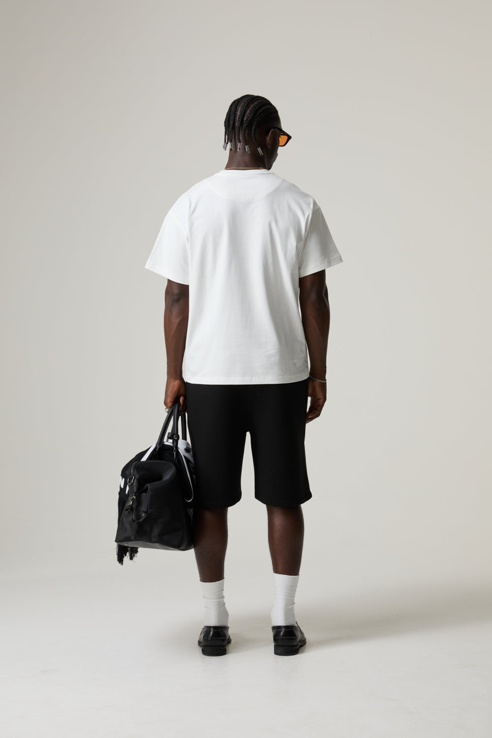 Model wearing a BOY LONDON white t-shirt and black shorts, holding a black bag on a plain background