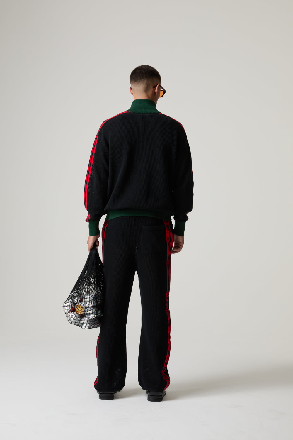 Model wearing a BOY LONDON black tracksuit with red stripes, holding a black bag with colourful design, on a white background.