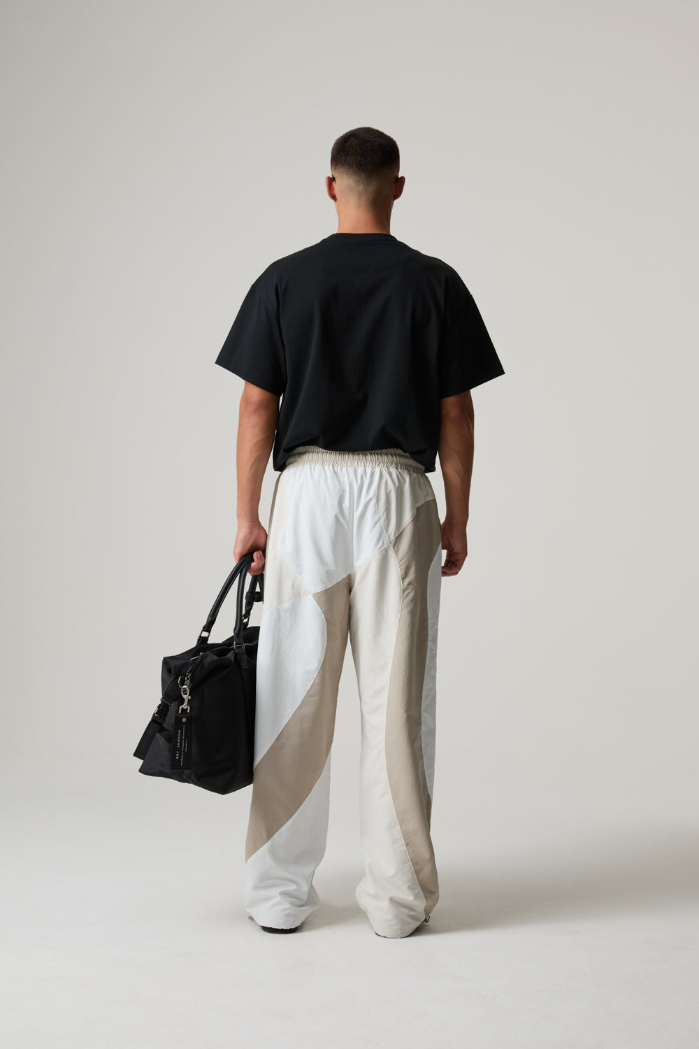 Model wearing a BOY LONDON black t-shirt and white pants holding a black bag on a plain background