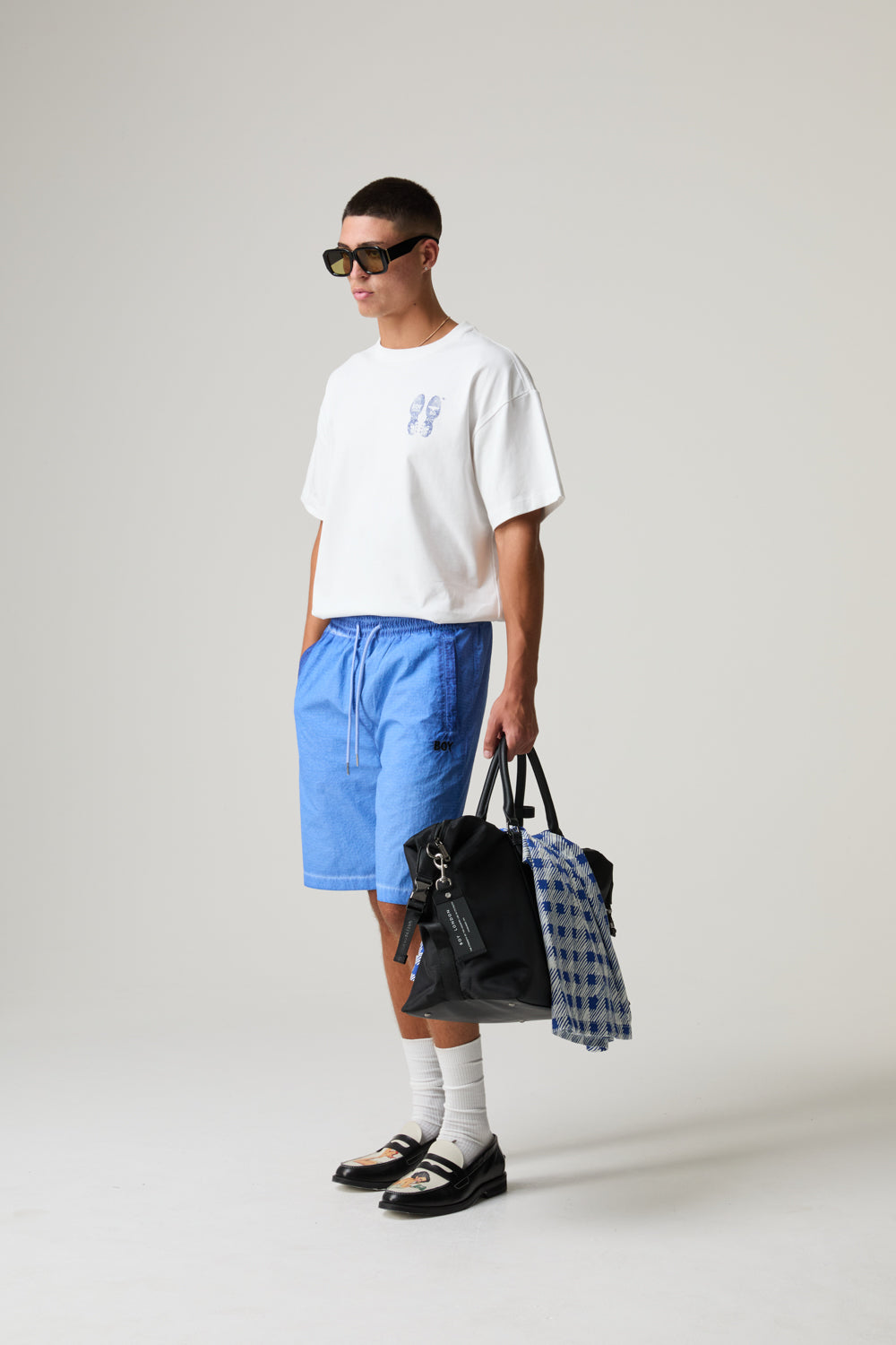 Model wearing a BOY LONDON white t-shirt, blue shorts, and black shoes holding bags against a plain background