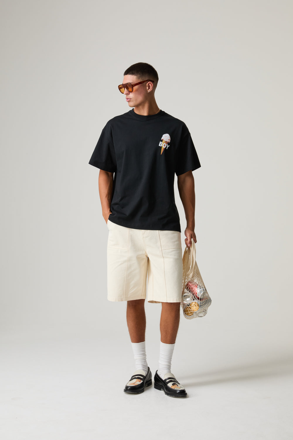 Model wearing a black BOY LONDON t-shirt with a graphic, beige shorts, white socks, and black sandals on a light gray background.