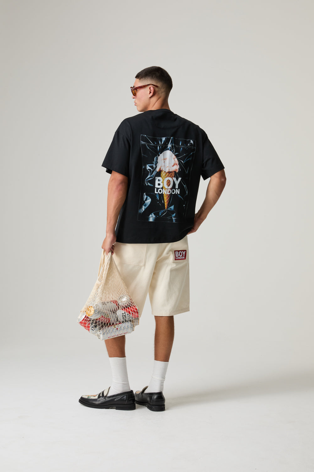 Model wearing a black BOY LONDON t-shirt with graphic design and text, beige shorts, and white sneakers on a plain background
