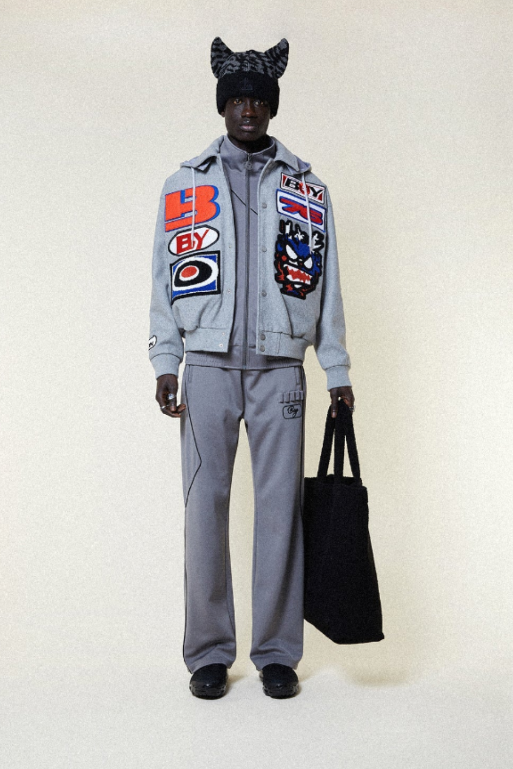 Person wearing a gray BOY London tracksuit with colorful patches and a black cat-themed hat, holding a black bag.