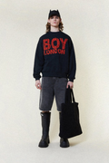 Model wearing a black 'BOY LONDON' sweatshirt with red text, holding a black bag on a white background.