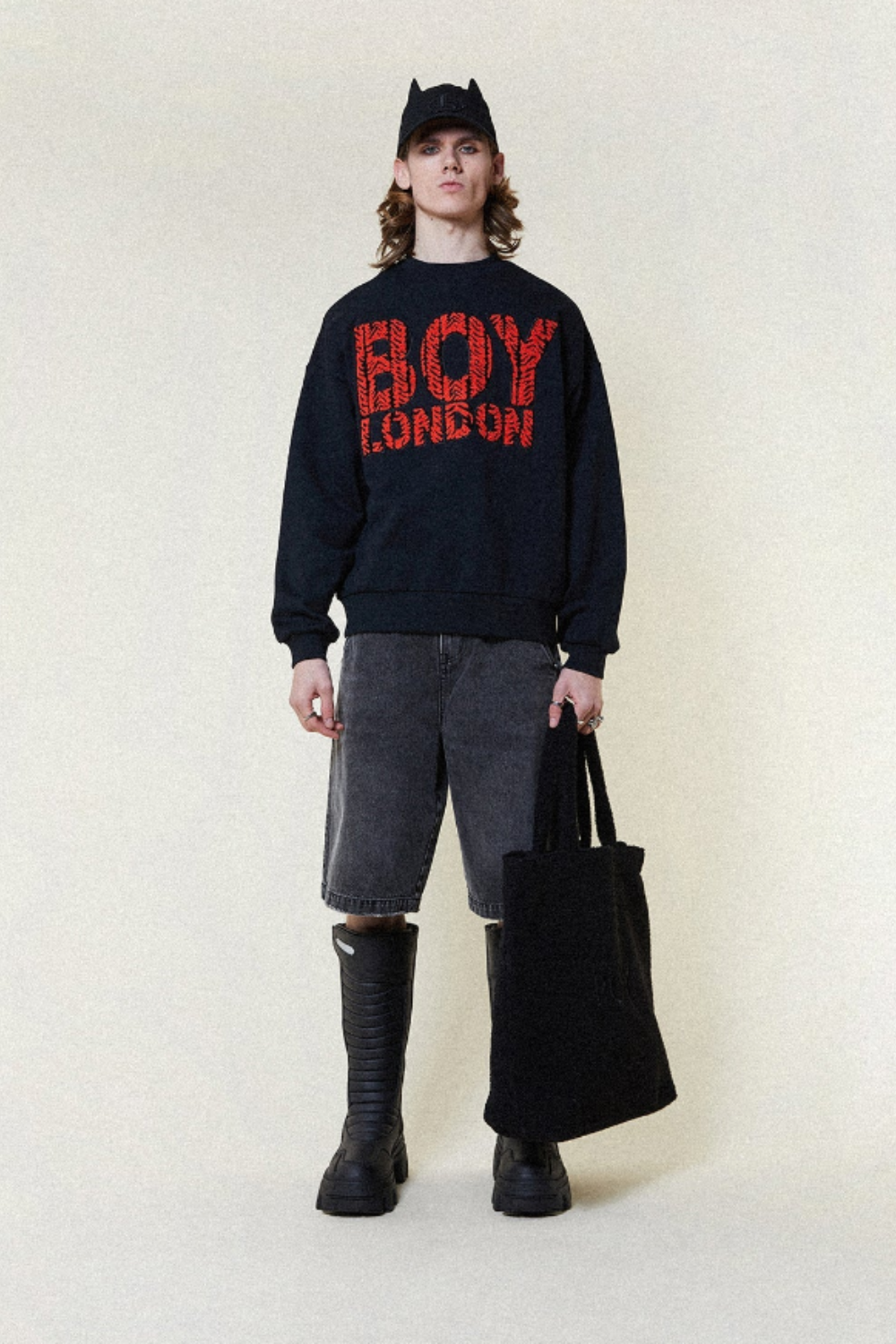 Model wearing a black 'BOY LONDON' sweatshirt with red text, holding a black bag on a white background.