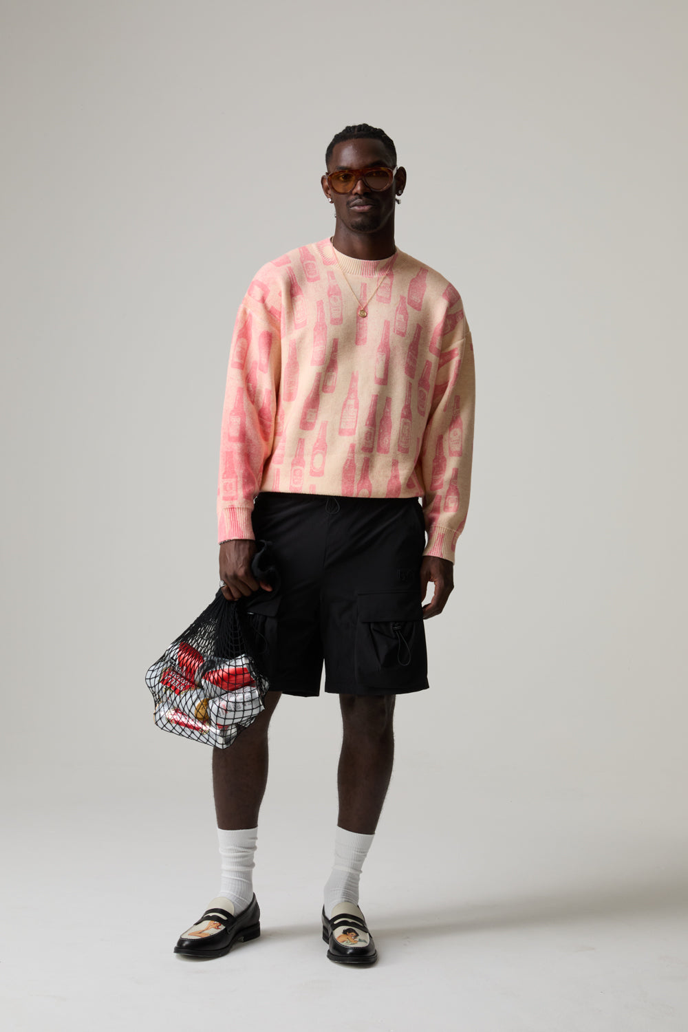 Model wearing a pink BOY LONDON sweater and black shorts, holding a patterned bag on a plain background