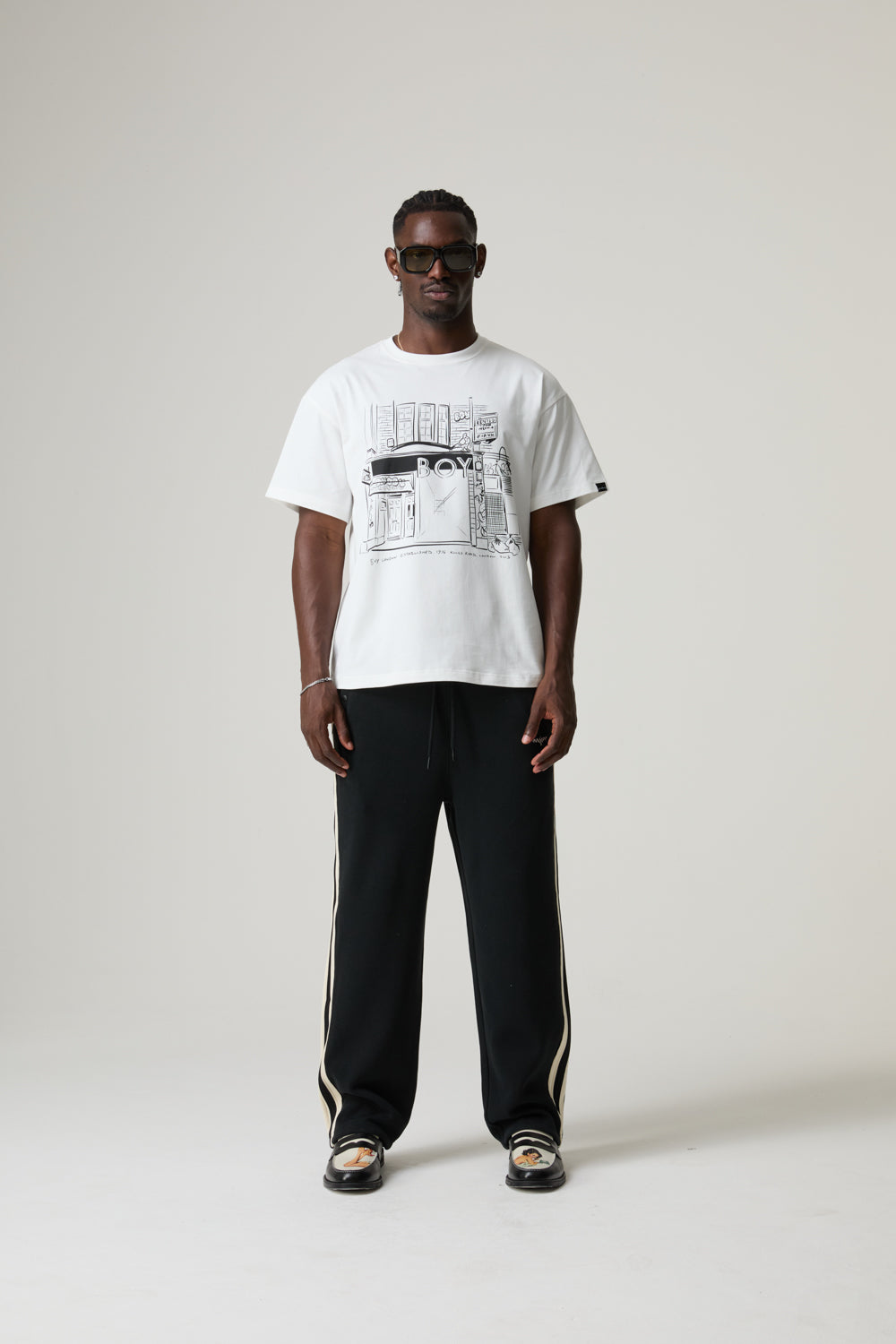 Model wearing a BOY LONDON white t-shirt with a graphic design and black pants on a plain background