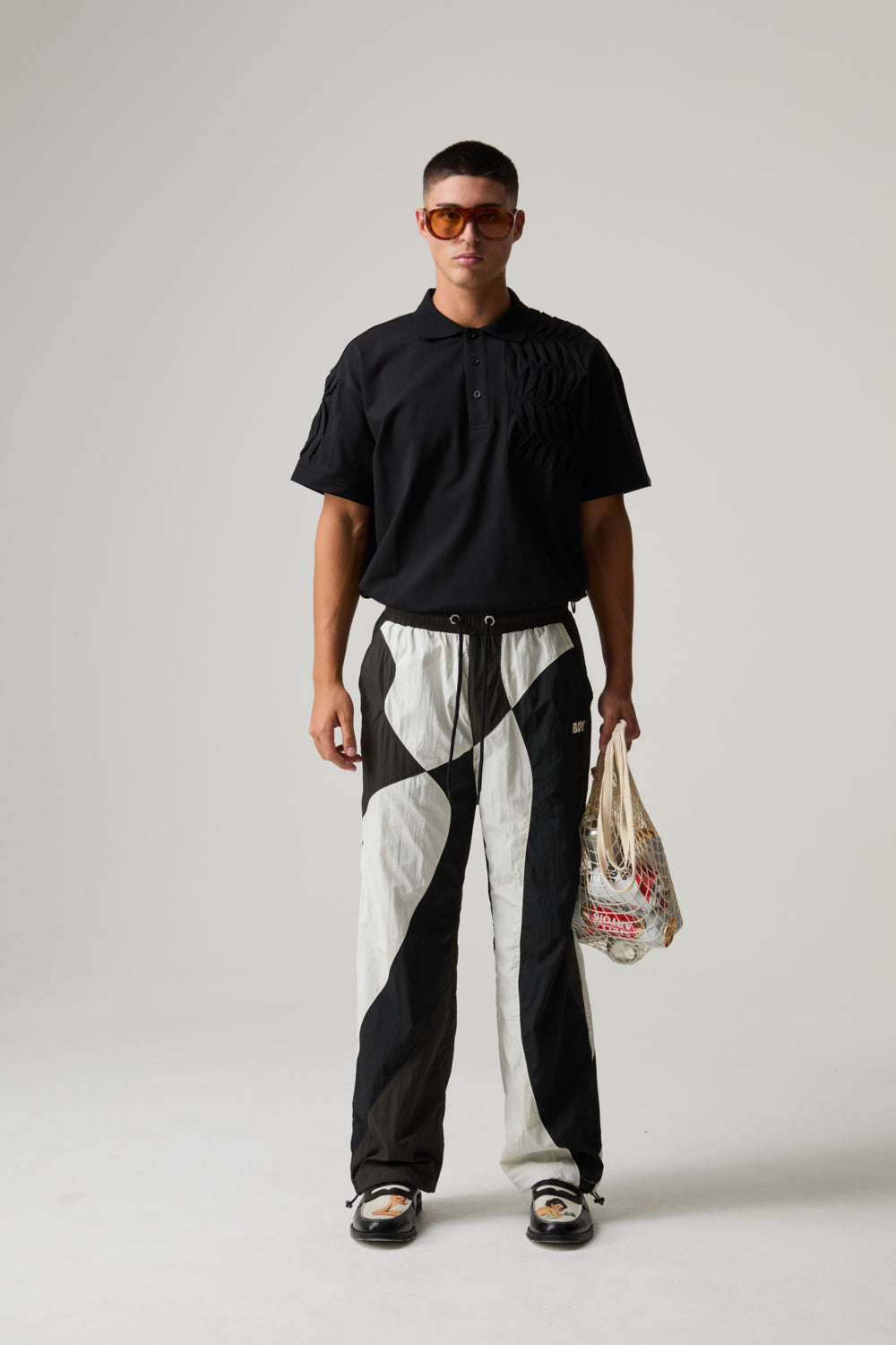 Model wearing a BOY LONDON black polo shirt and black and white pants, holding a bag.