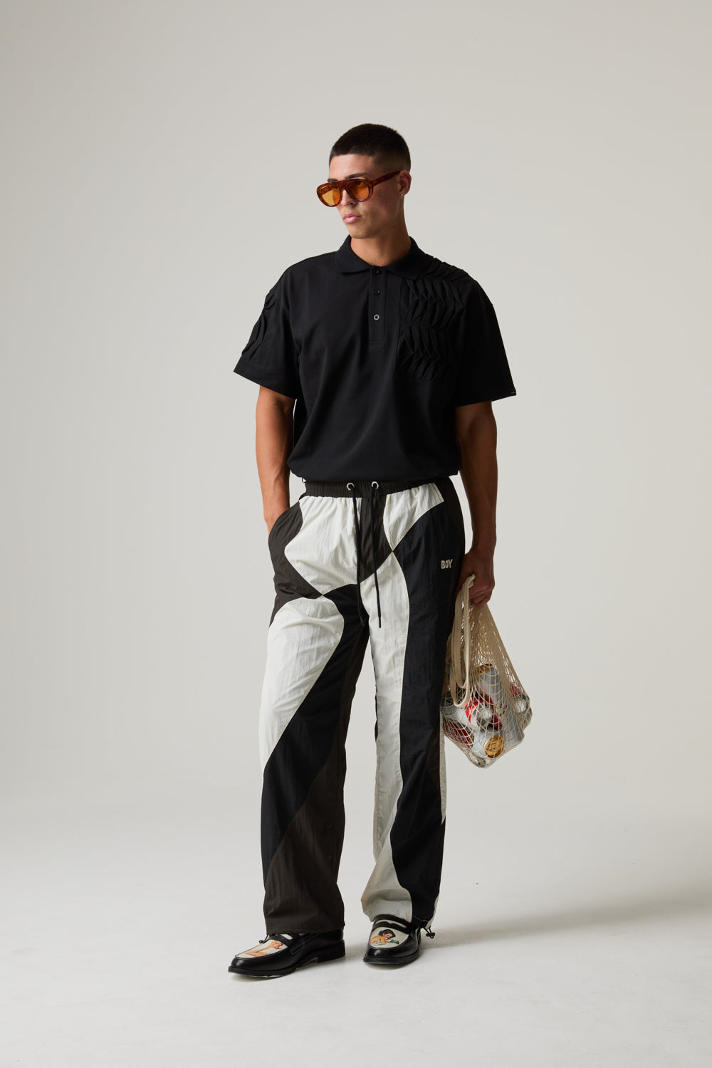 Model wearing a black BOY LONDON polo shirt and black and white pants holding a plastic bag on a plain background