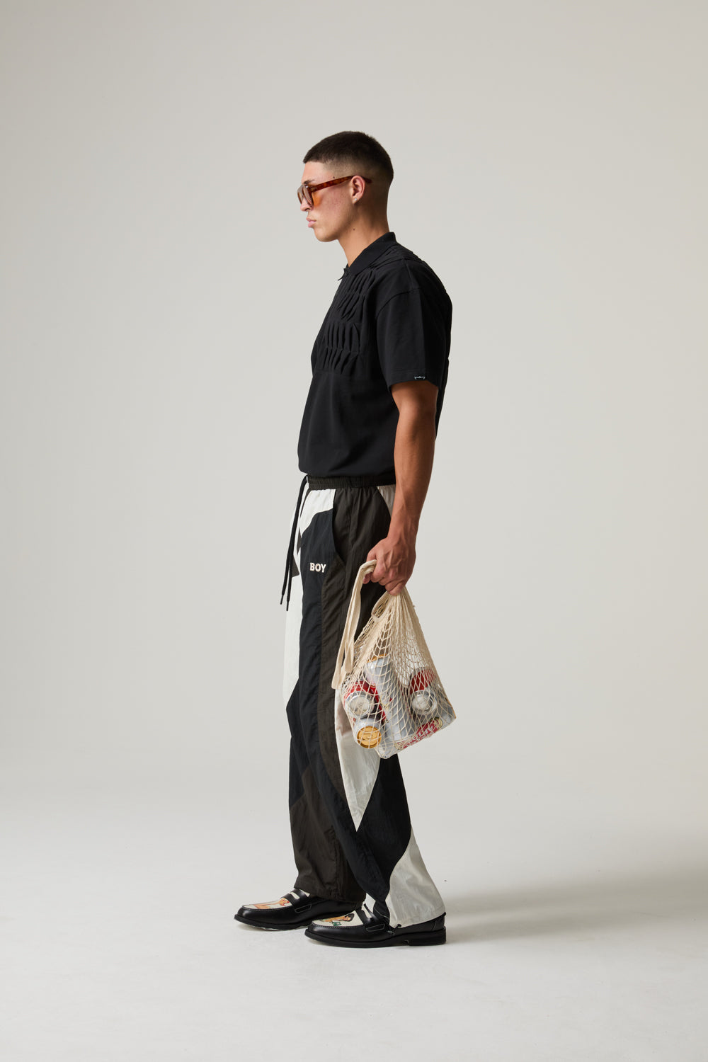 Model wearing a black BOY LONDON outfit with a white apron holding a patterned bag on a plain background