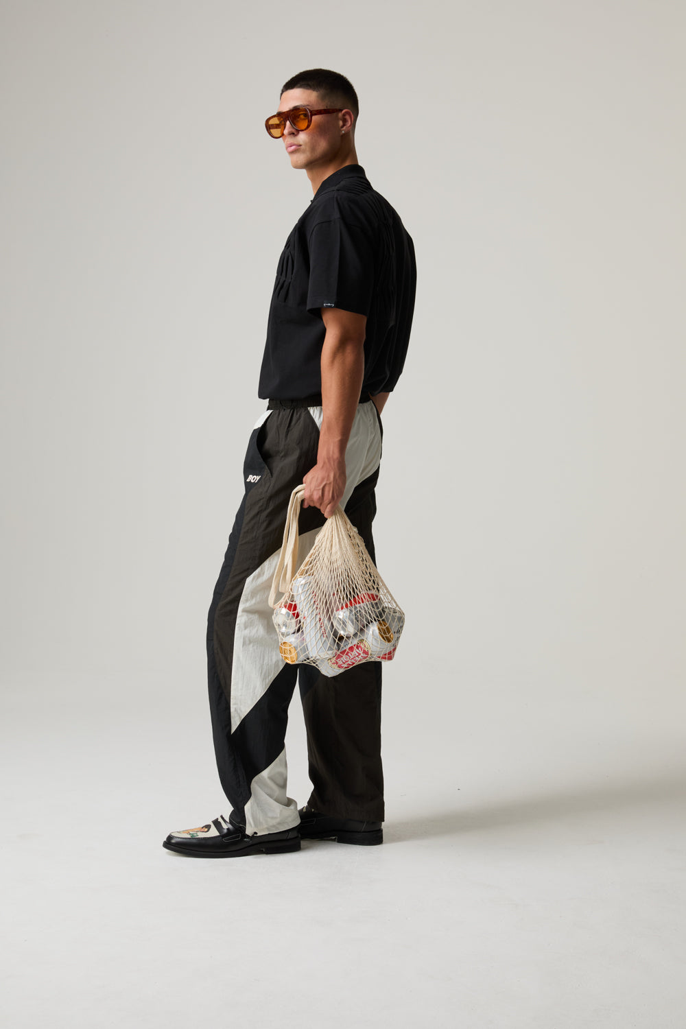 Model wearing a BOY LONDON outfit, holding a plastic bag with a pattern, wearing sunglasses and black shoes on a plain background