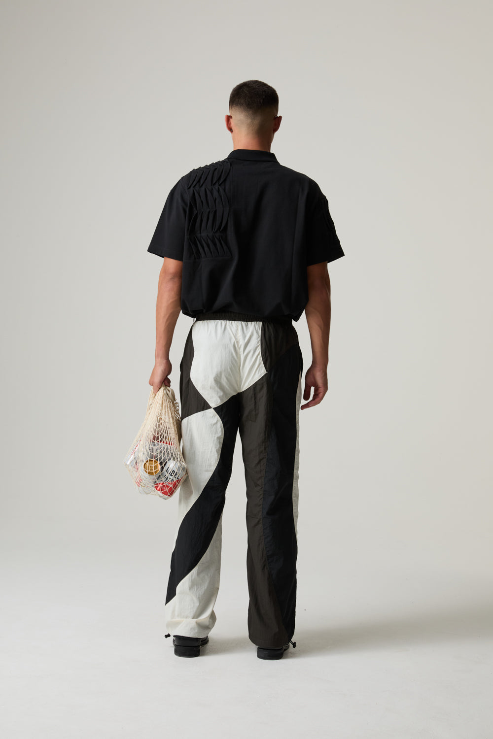 Model wearing a black BOY LONDON shirt and white pants with black stripes, holding a colourful bag.