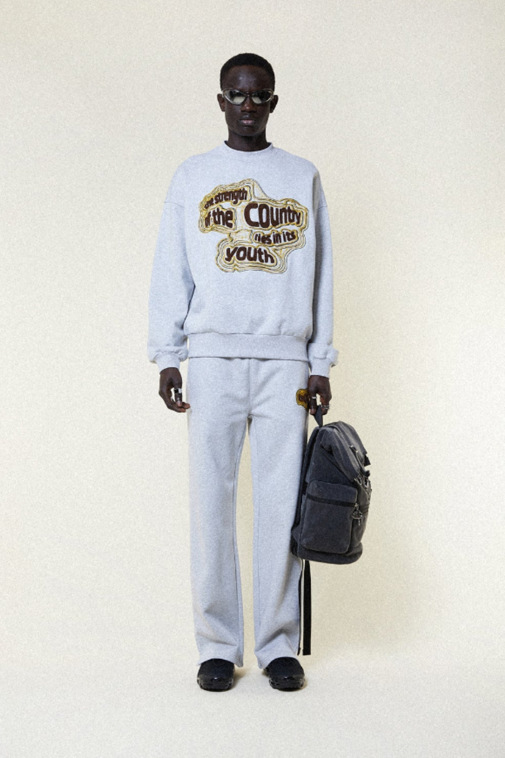 Male model wearing a light blue BOY London sweatshirt with text and holding a black bag on a white background