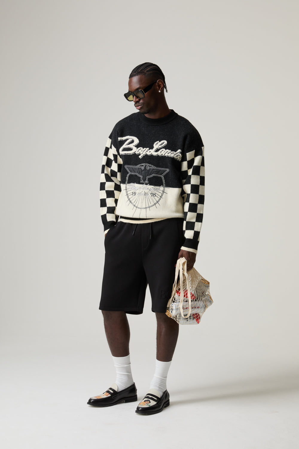 Model wearing a black and white BOY LONDON checkered sweater with text, black shorts, and holding a bag on a plain background