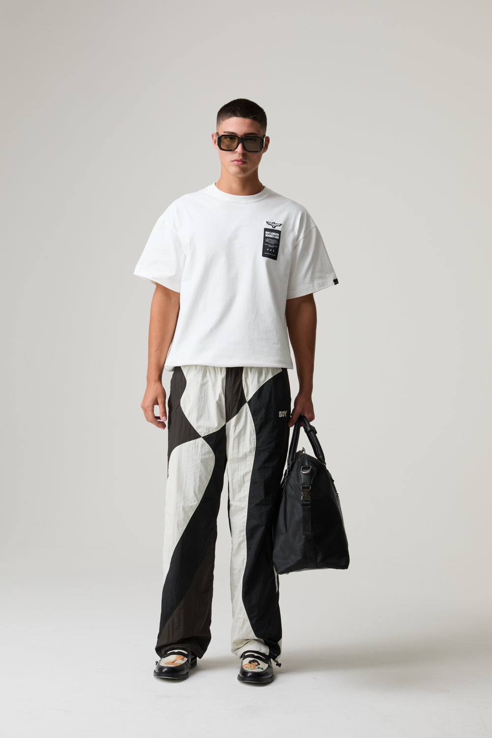 Model wearing a BOY LONDON white t-shirt with a black graphic, black and white pants, and sunglasses, holding a black bag on a plain background.