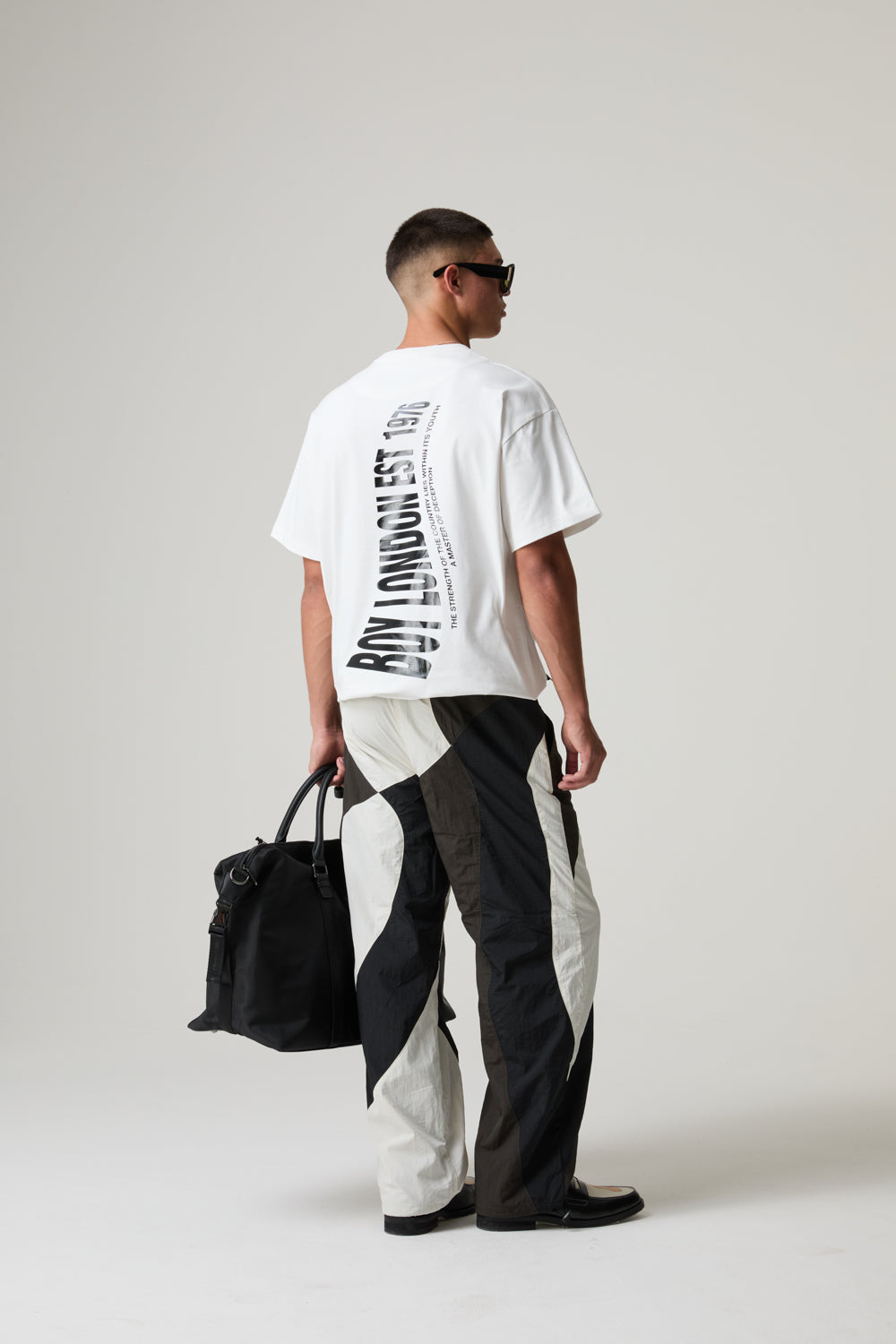 Model wearing a BOY LONDON white t-shirt with text and black and white pants, holding a black bag on a plain background
