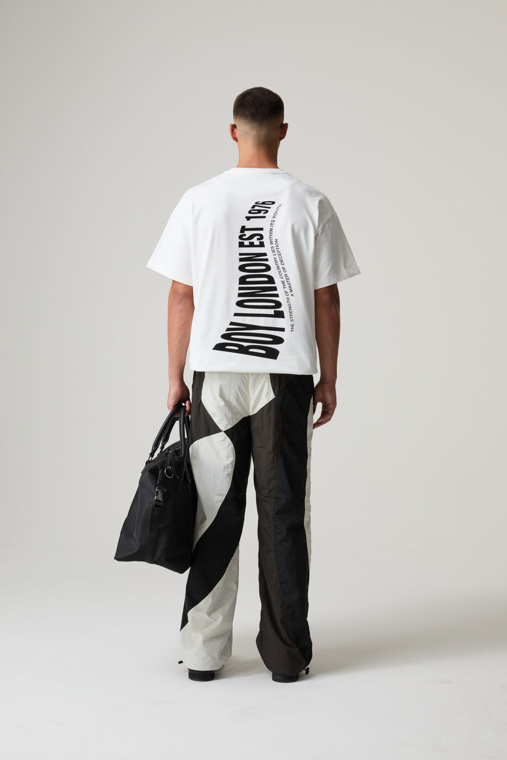 Model wearing a BOY LONDON white t-shirt with text and black and white pants, holding a black bag on a plain background